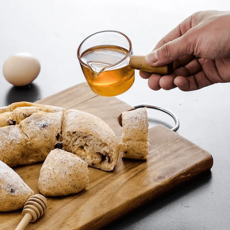 100 ml Glass Espresso Measuring Cup with wooden handle used to pour syrup over bread, perfect for accurate liquid measurement in kitchen tasks.