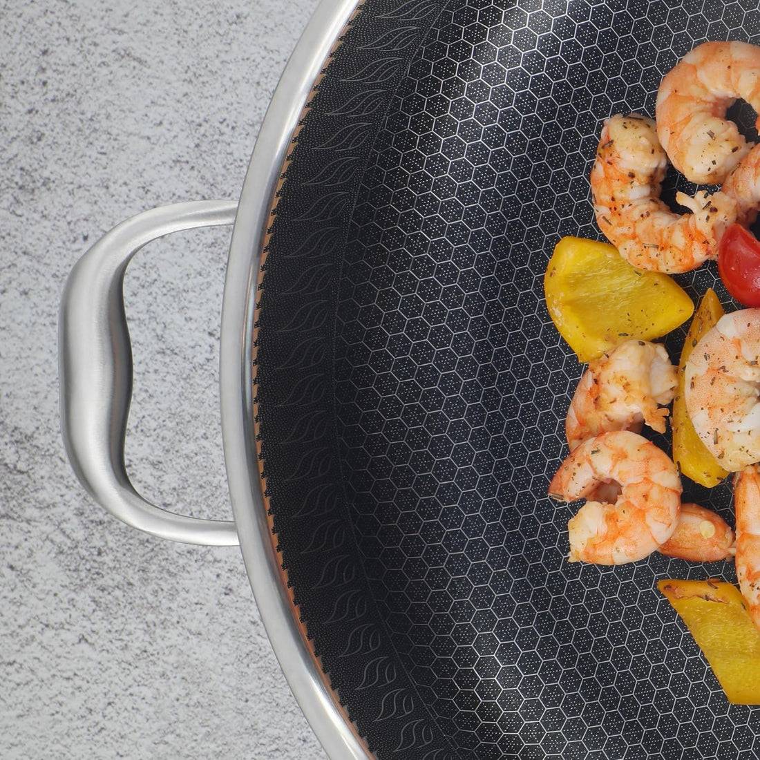 Close-up of a stainless steel non-stick frying pan with two handles, showcasing its hexagonal textured surface while cooking shrimp and vegetables.